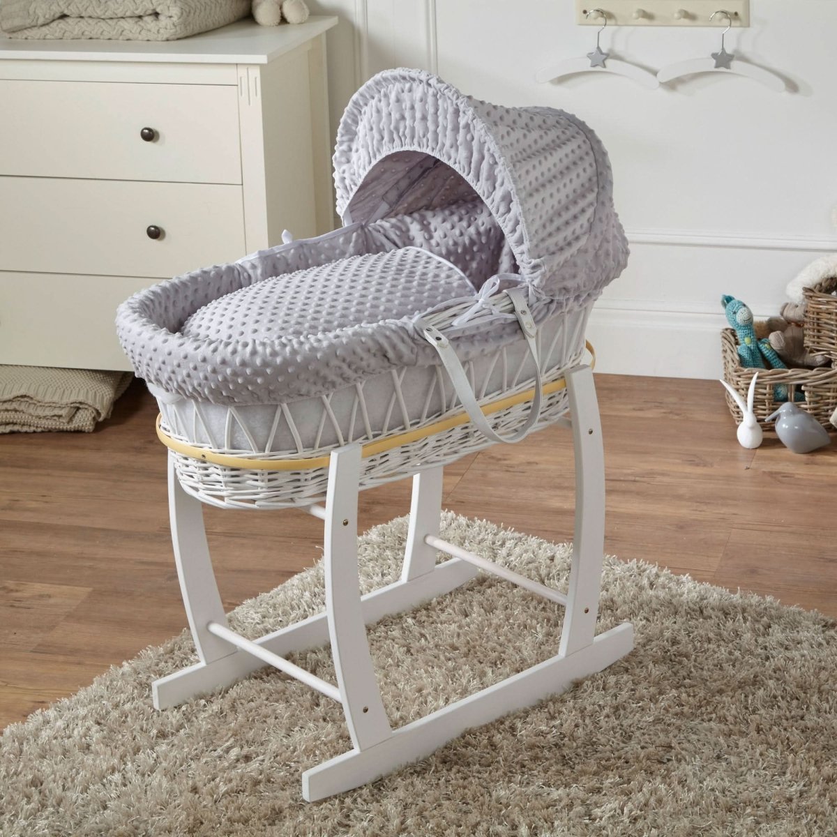 Gray wicker Moses basket with hood on a white stand in a nursery setting.